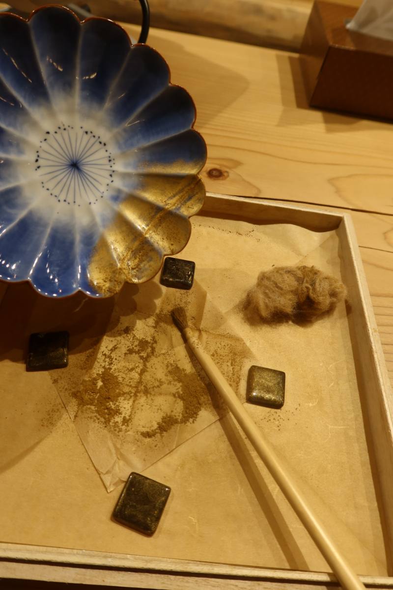 Gold powder and a fine brush on a craft workbench, beside a porcelain dish